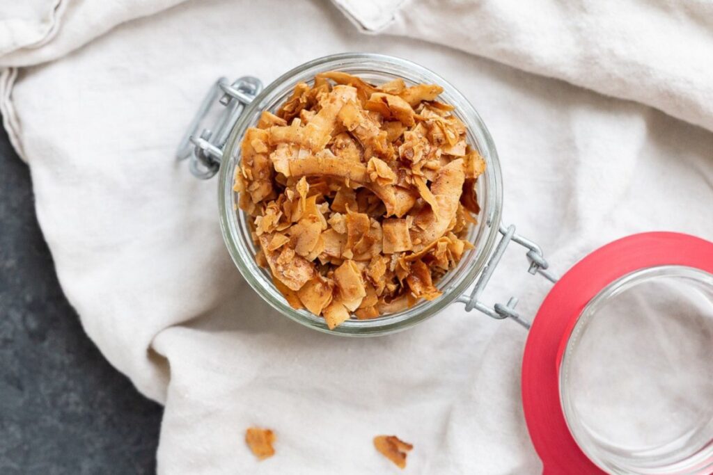 Top view of a jar filled with toasted coconut flakes, placed on a light-colored cloth. Some coconut flakes are scattered around the jar on the fabric. A lid with a red rim is partially visible to the side.