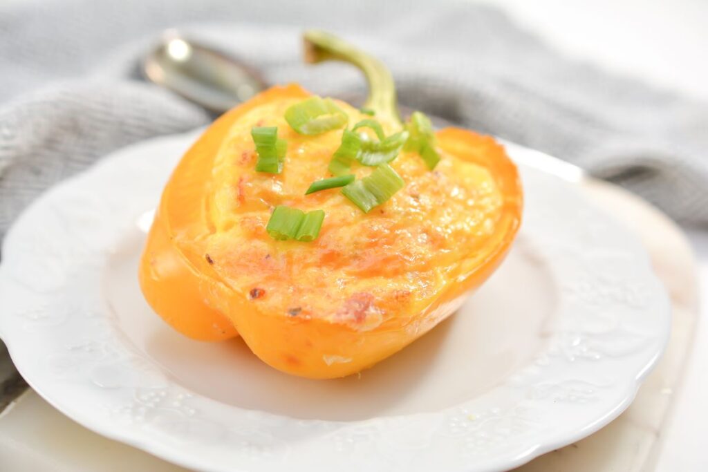 A baked half of a yellow bell pepper filled with a cheesy mixture, topped with sliced green onions, on a white plate with a grey cloth in the background.