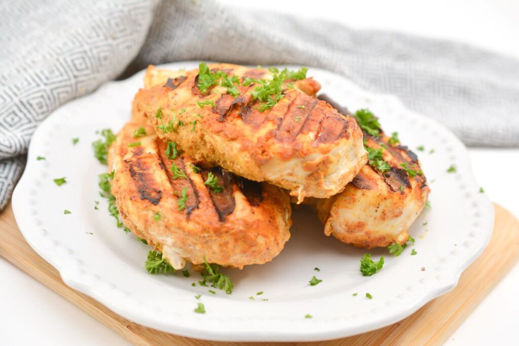 Grilled, seasoned chicken breasts garnished with chopped parsley on a white plate. A gray patterned cloth is seen in the background.