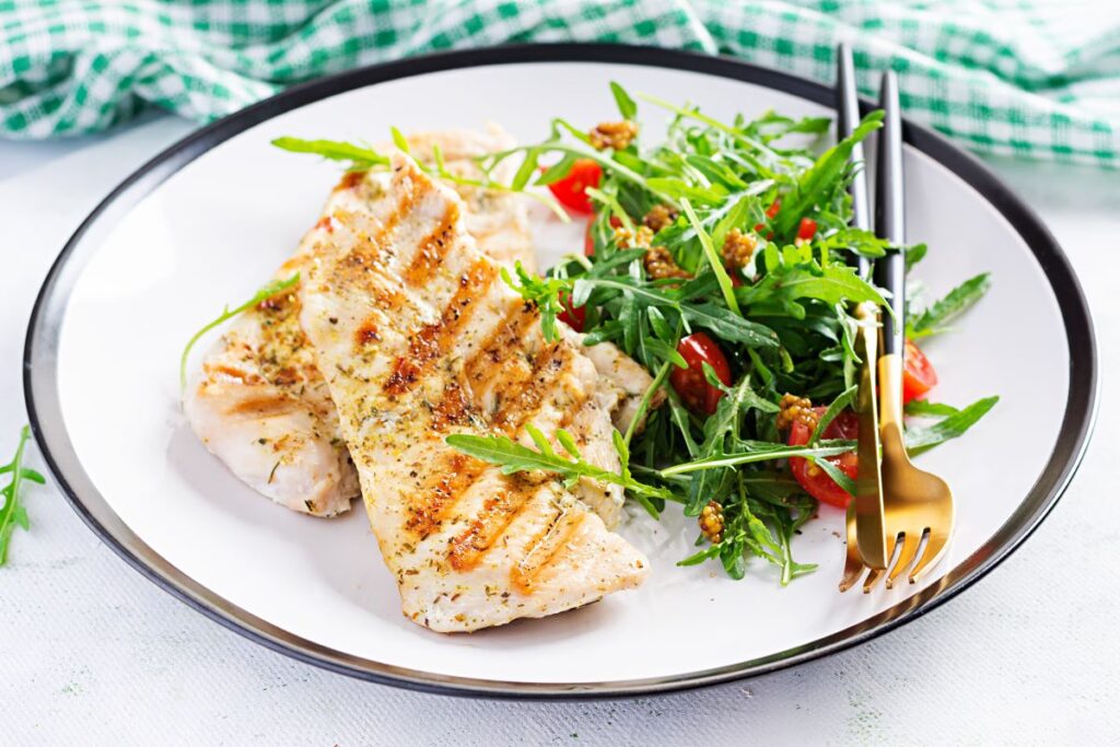 Grilled chicken breast served with a fresh salad of arugula, cherry tomatoes, and nuts on a white plate. A fork and knife are placed beside the salad, with a green checkered cloth in the background.