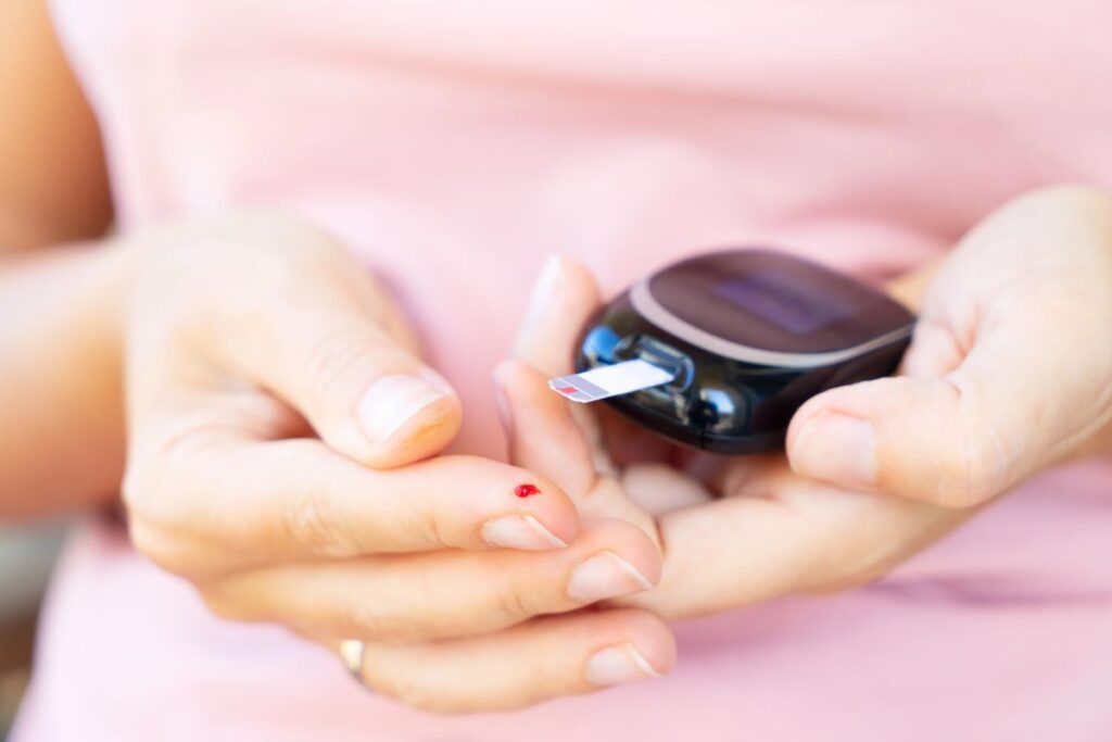 A person holding a blood glucose meter with a test strip inserted. A small drop of blood is visible on their fingertip, indicating they are checking their blood sugar level. They are wearing a light pink shirt.