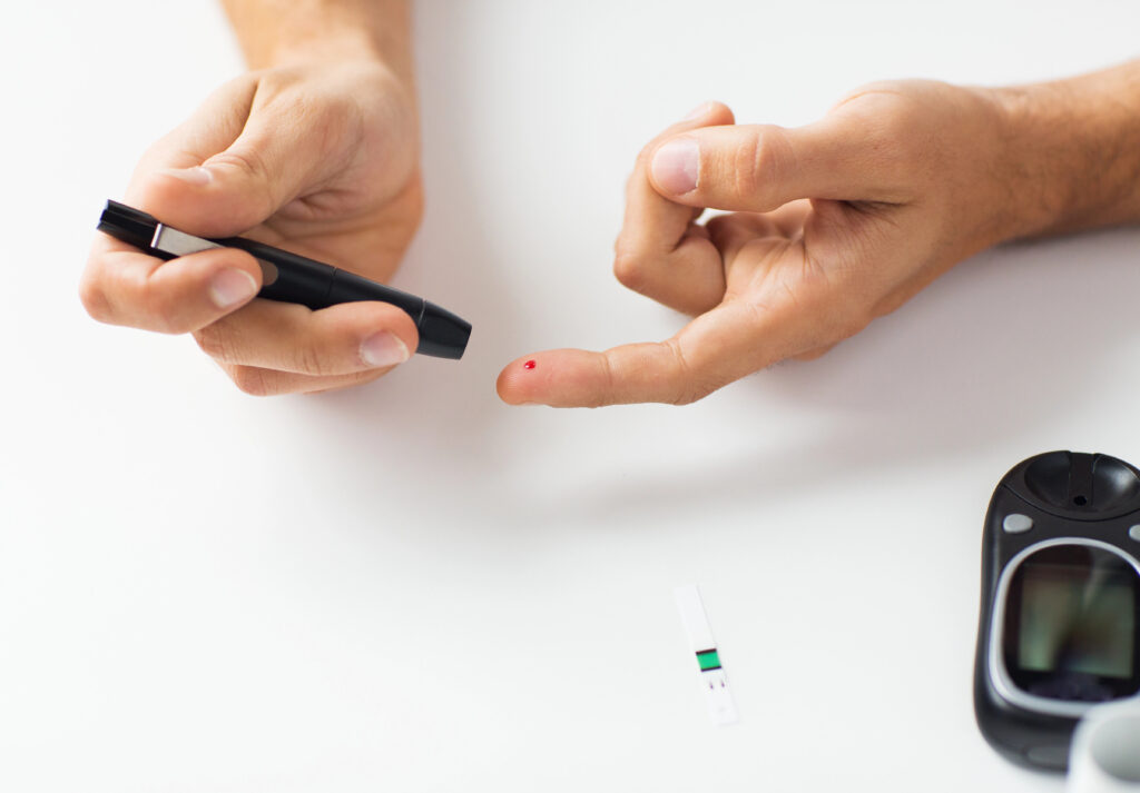 A person uses a lancet on their fingertip for a blood sugar test. A small drop of blood is visible on the finger. A glucose meter and test strip are on the white surface nearby.