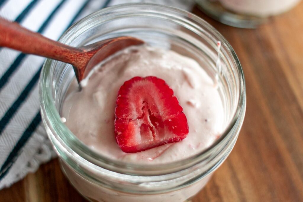 A glass jar filled with creamy pink yogurt sits on a wooden surface. A wooden spoon rests inside the jar, and a fresh strawberry slice decorates the top. A striped cloth is partially visible beside the jar.